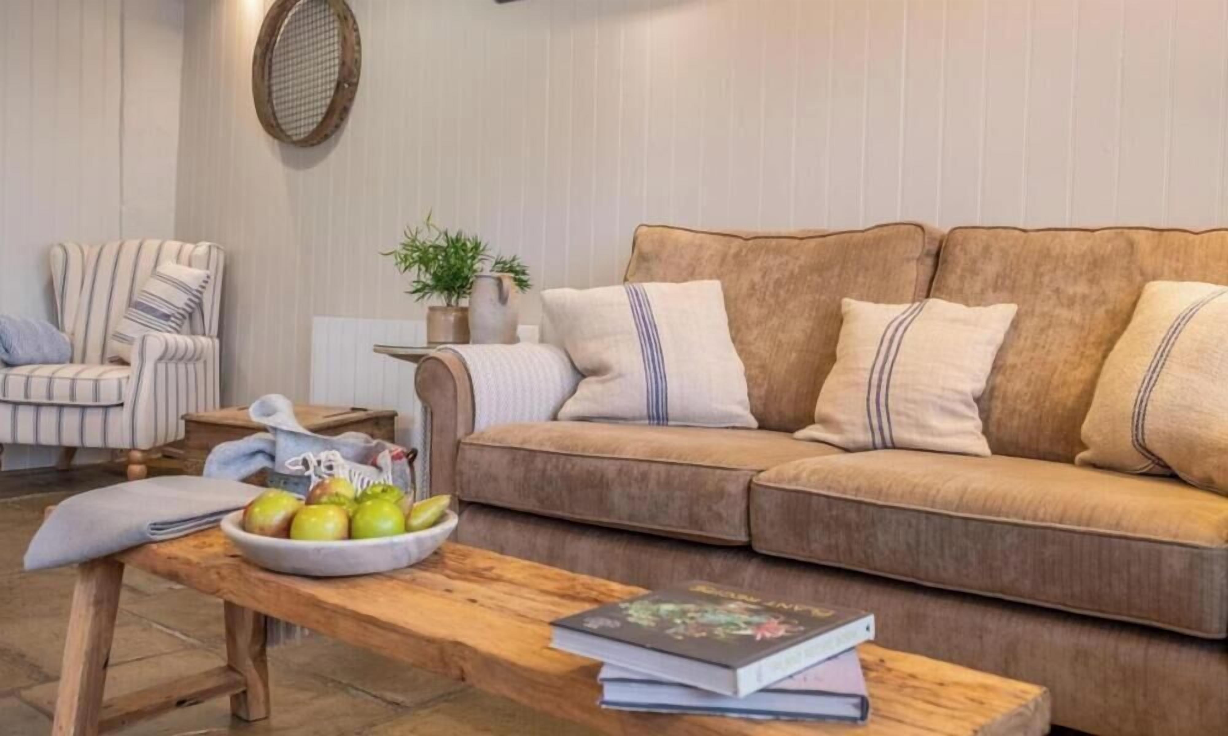 Ein gemütliches Wohnzimmer mit einem braunen Sofa, einem gestreiften Sessel, einem Holztisch mit Obst und Büchern sowie einer Zimmerpflanze.