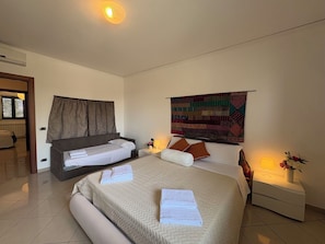 Une chambre d’hôtel comprenant un lit, un petit lit, une table de chevet, une lampe, une tapisserie murale et une fenêtre avec des stores.