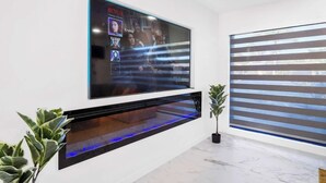 Een moderne woonkamer met een grote flatscreen televisie aan de muur, een ingebouwde open haard eronder en een potplant aan de rechterkant.