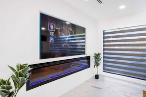Ein modernes Wohnzimmer mit einem großen Flachbildfernseher an der Wand, darunter ein eingebauter Kamin und rechts davon ein Topfpflanze.