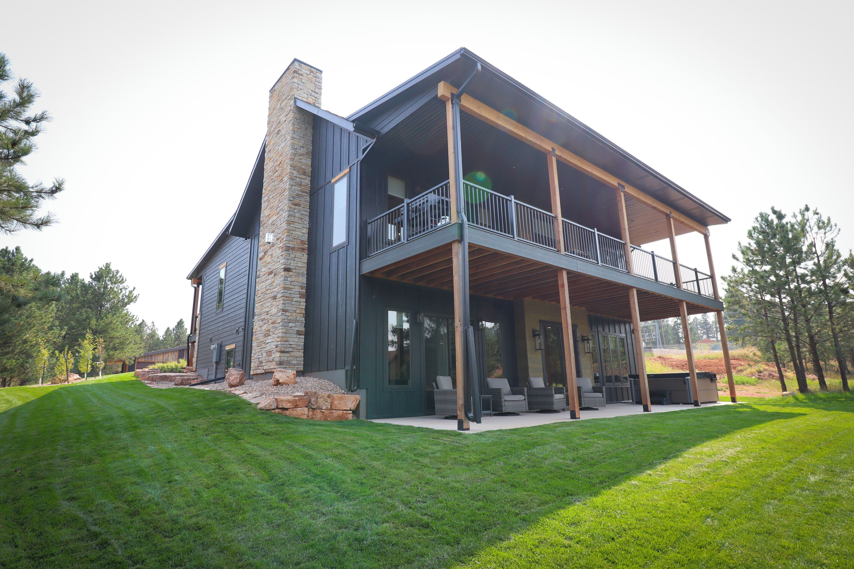 A modern two-story house with a stone chimney, a balcony with outdoor furniture, and a well-maintained lawn.