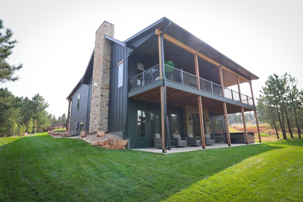 Une maison moderne à deux étages, dotée d’une cheminée en pierre, d’un balcon avec des meubles d’extérieur et d’une pelouse bien entretenue.