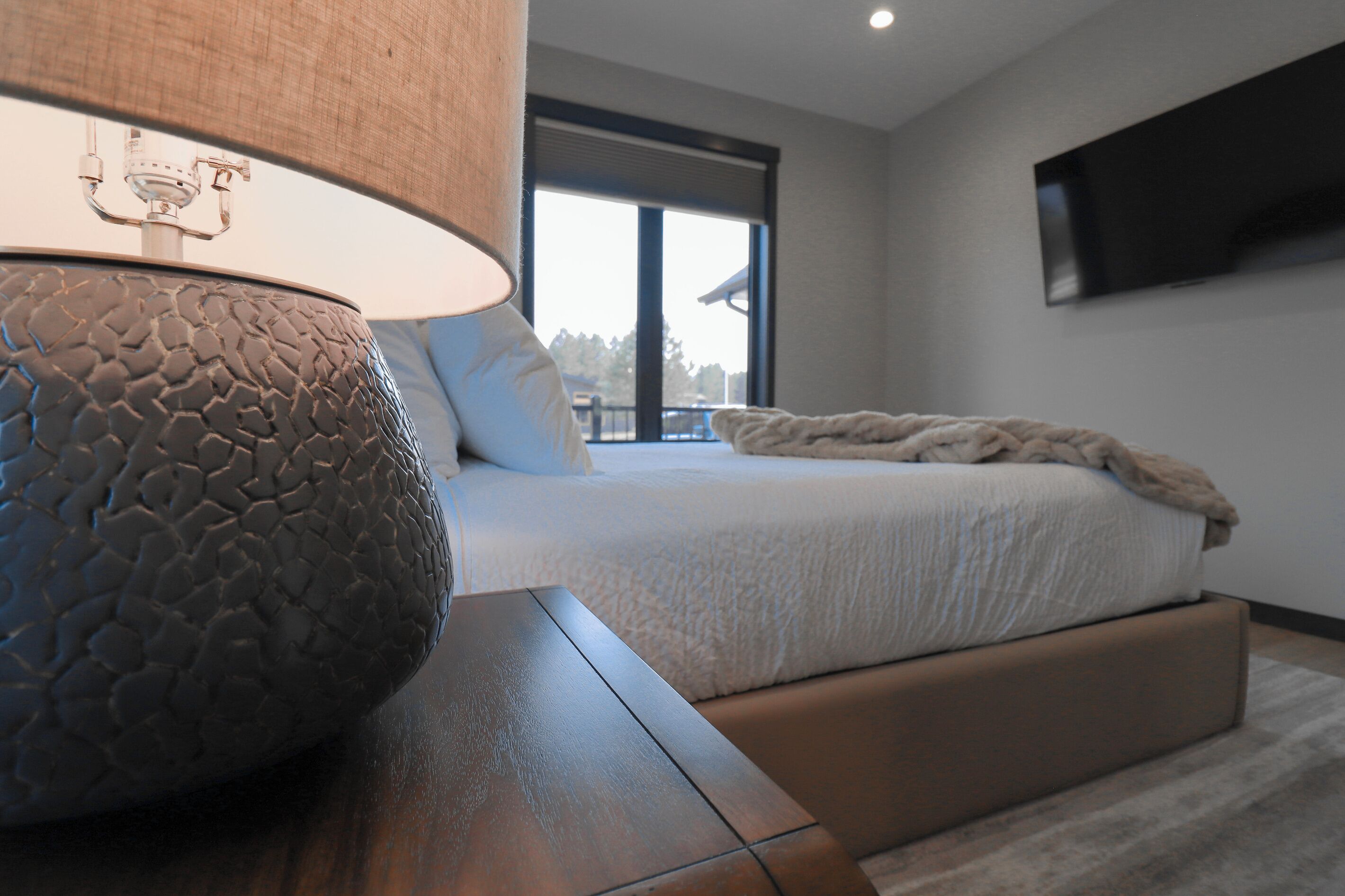 A modern bedroom with a large bed, a bedside table with a textured lamp, and a TV mounted on the wall.