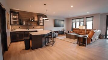 Una cocina moderna con una isla central, un comedor con sofá y mesa de centro, y una sala de estar con un ventanal grande.
