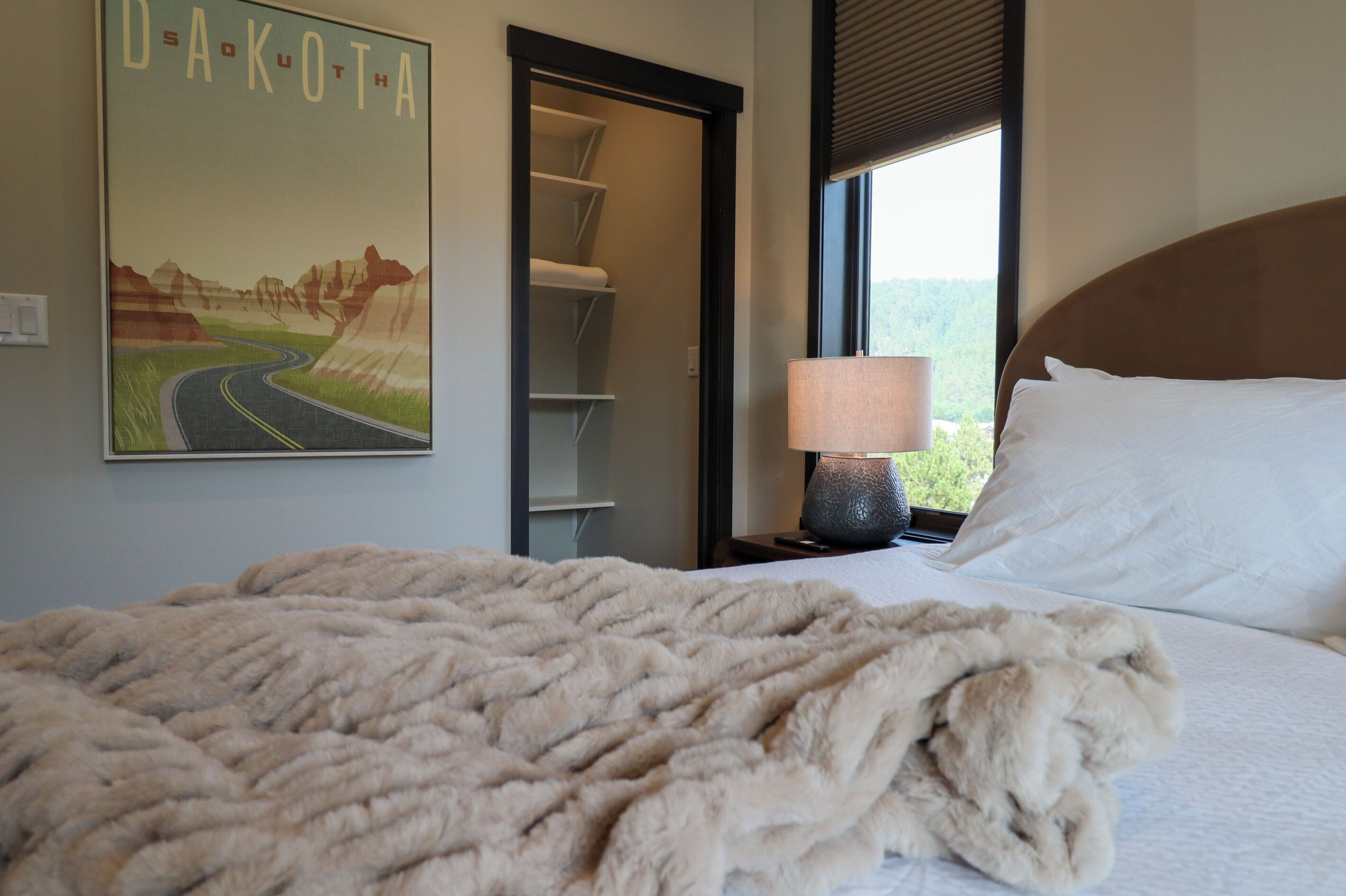 Una habitación de hotel con una cama, una mesita de noche con lámpara, una ventana con vistas y una puerta que da a un armario.