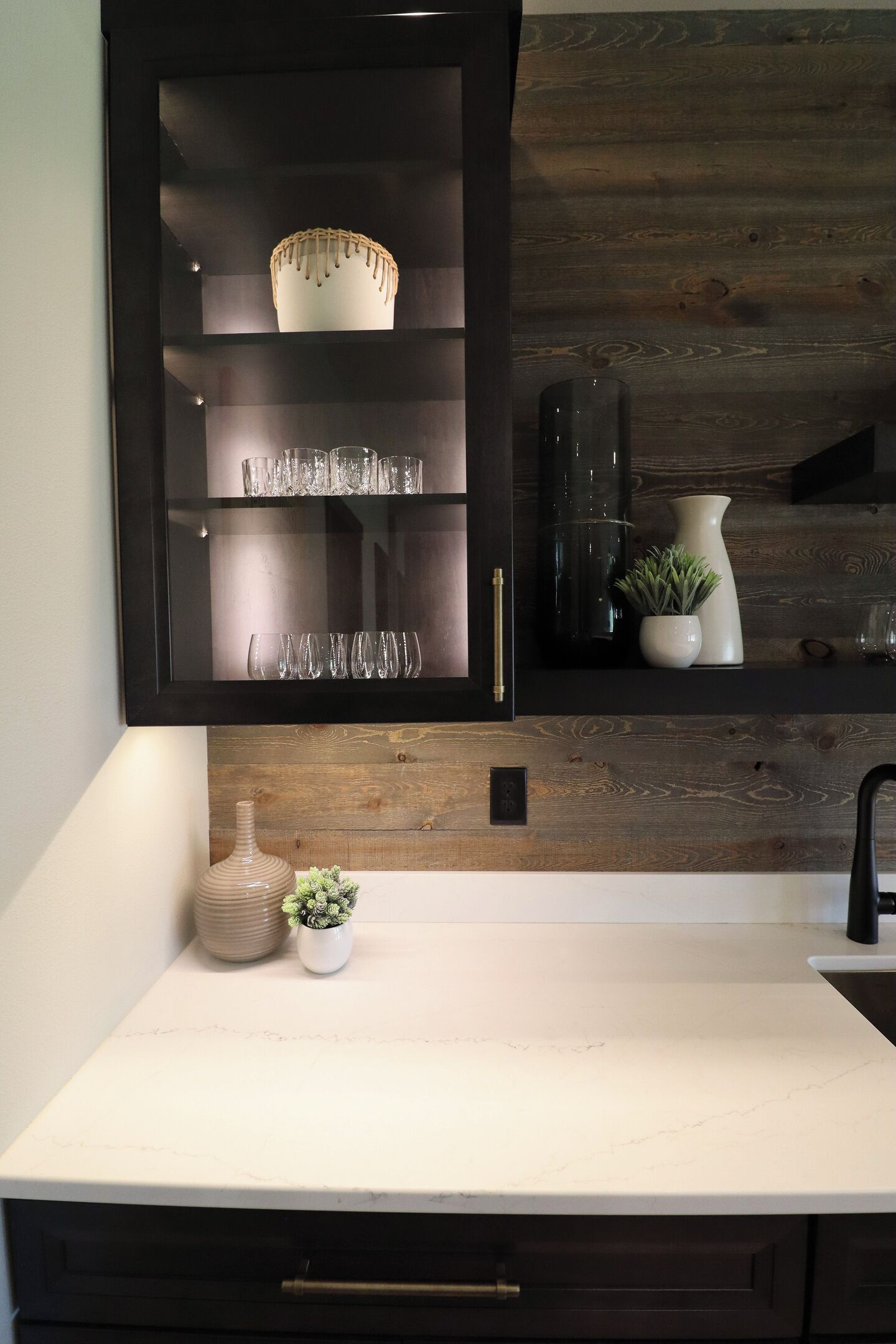 A kitchen with a dark wooden cabinet, a white countertop, and a rustic wooden wall.