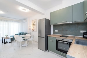 A modern kitchen with a built-in oven, a refrigerator, and a dining area with a table and chairs. - Athina Residential Complex (Paralia Ofriniou)