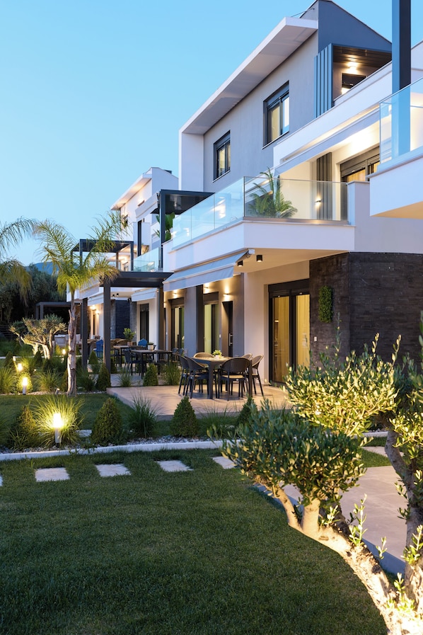 A modern multi-story residential building with a well-maintained garden and outdoor seating area. - Athina Residential Complex (Paralia Ofriniou)