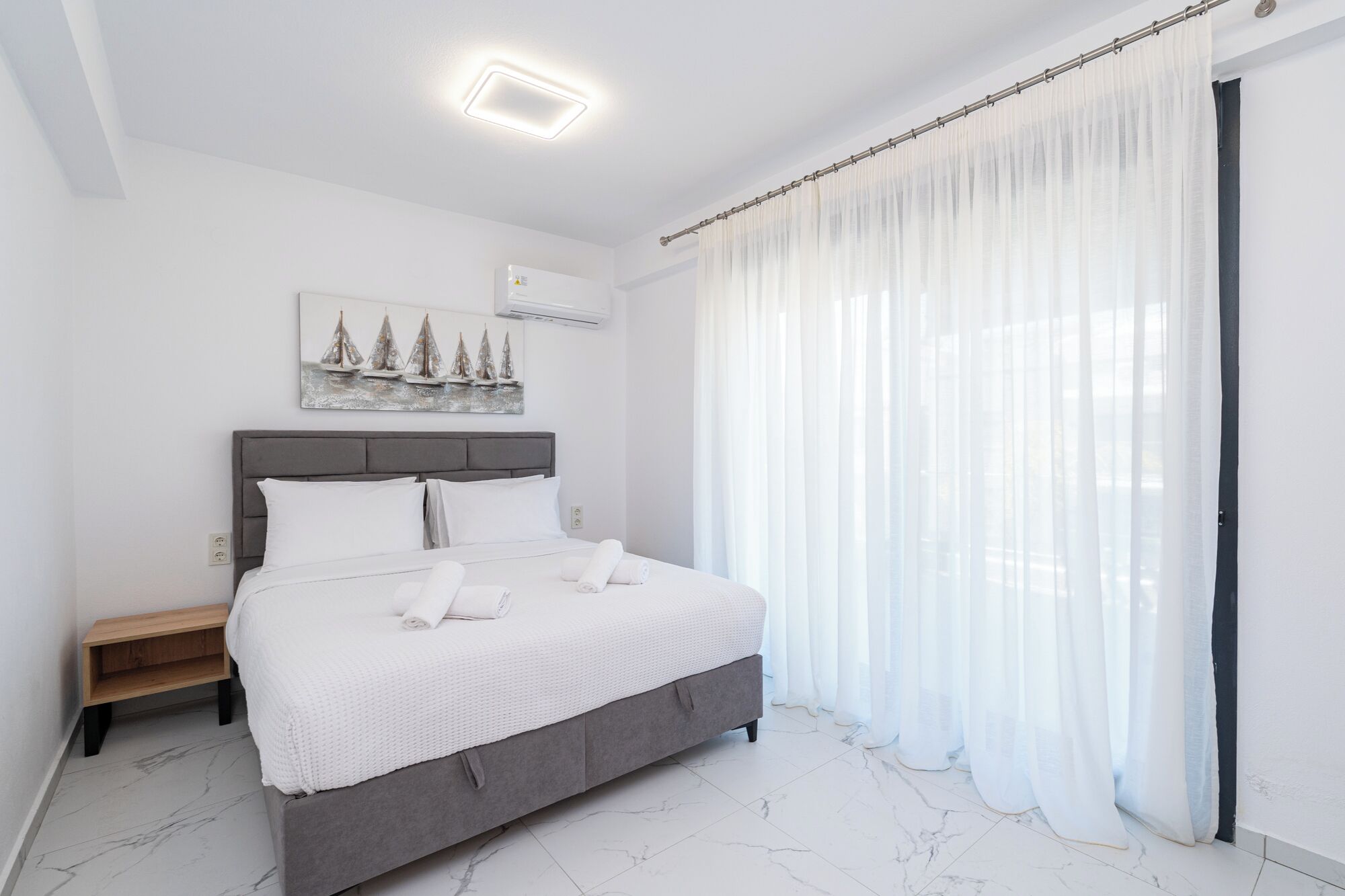A bedroom with a bed, a nightstand, a painting, a window with sheer curtains, and a ceiling light.