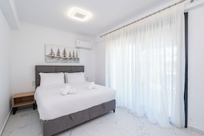 A bedroom with a bed, a nightstand, a painting, a window with sheer curtains, and a ceiling light. - Athina Residential Complex (Paralia Ofriniou)