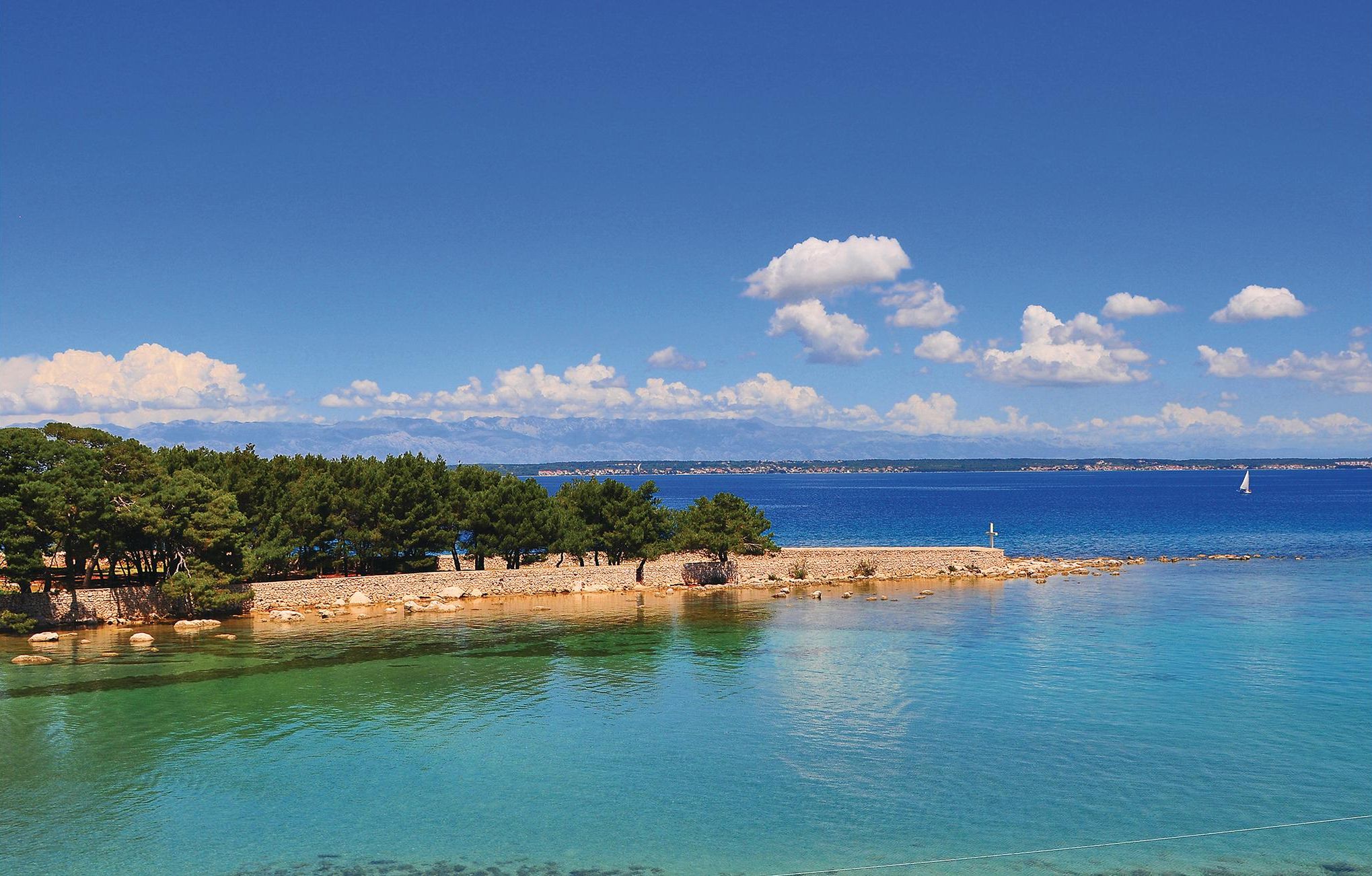 A coastal landscape with clear turquoise waters, a rocky shoreline, and a backdrop of mountains and a clear sky.