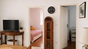 Une pièce avec une télévision posée sur une table en bois, une horloge au mur et un lit recouvert d’une couverture aux couleurs vives.