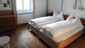 En dobbeltseng med hvide sengetøj, en trækommode, et lille bord og et vindue med gardiner.