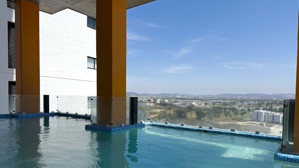 An indoor infinity pool with clear blue water, surrounded by beige pillars, and a view of a cityscape. - Accommodation with beautiful views, with a pool. (Mazatlán)