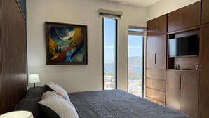 A bedroom with a large bed, a painting, a TV, and a view of the city. - Accommodation with beautiful views, with a pool. (Mazatlán)