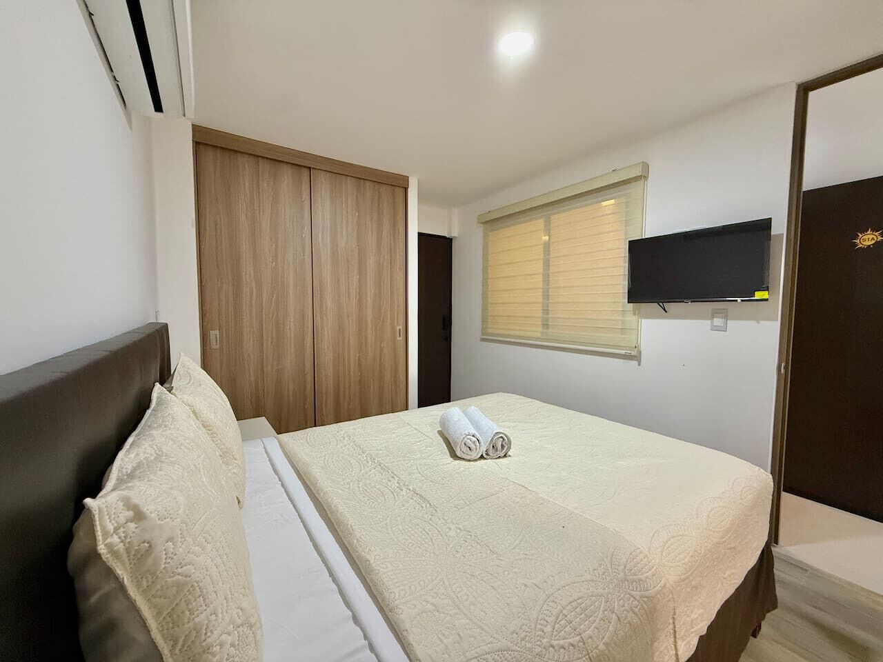 A hotel room with a bed, a wooden wardrobe, a television on the wall, and a window with blinds.