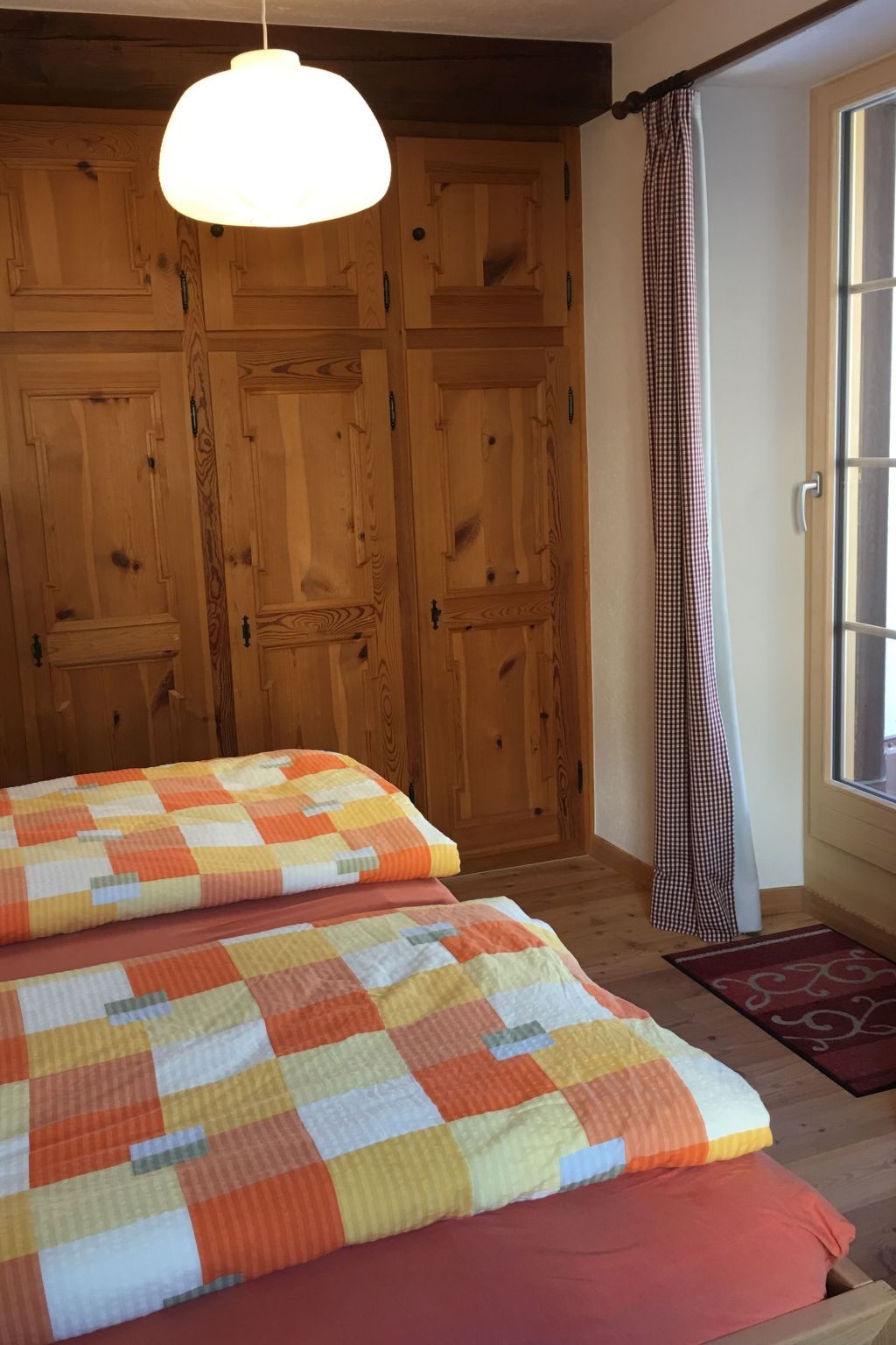 A bedroom with a wooden wardrobe, a checkered bedspread, and a window with curtains.