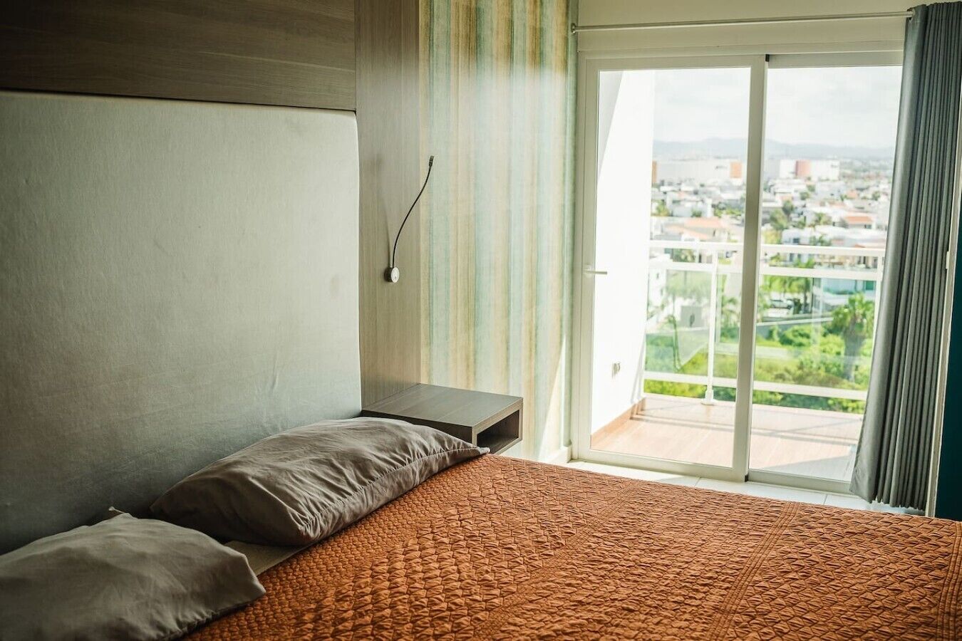 A hotel room with a bed, a bedside table, a wall-mounted phone, and a view of a cityscape through a large window.