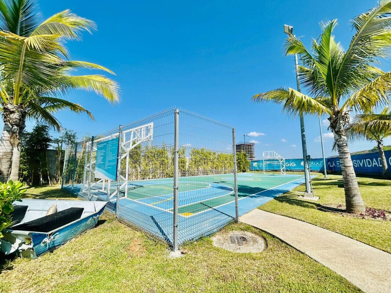 Een omheind sportterrein met een basketbalring, omgeven door palmbomen en een blauwe lucht.