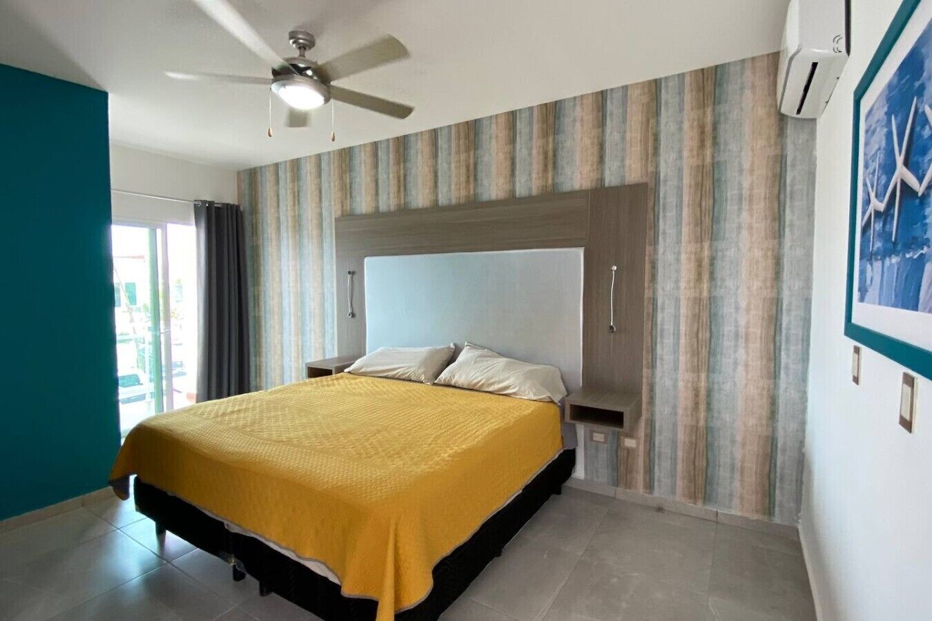 A hotel room with a bed, a ceiling fan, a window with curtains, and a wall-mounted air conditioner.
