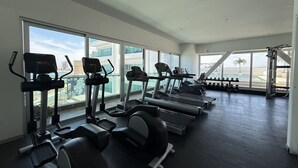 A gym with treadmills, elliptical machines, and dumbbells, large windows, and a view of buildings. - Departamento Premium Torre M by GPS (Mazatlán)