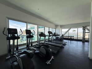 A gym with treadmills, elliptical machines, and dumbbells, large windows, and a view of buildings. - Departamento Premium Torre M by GPS (Mazatlán)