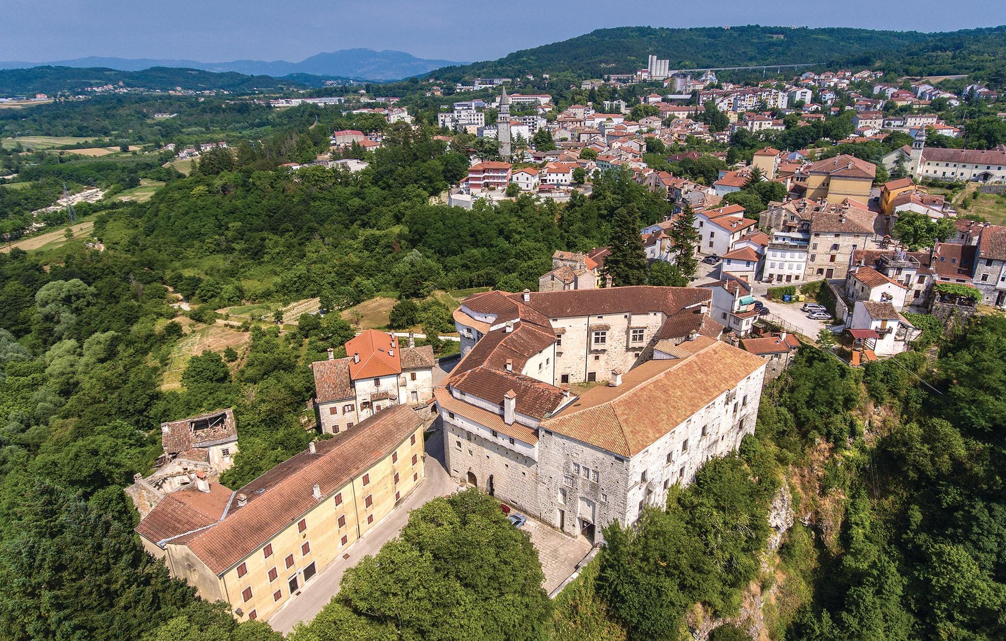 An aerial view of a historic town with stone buildings, a prominent church tower, and surrounding greenery.