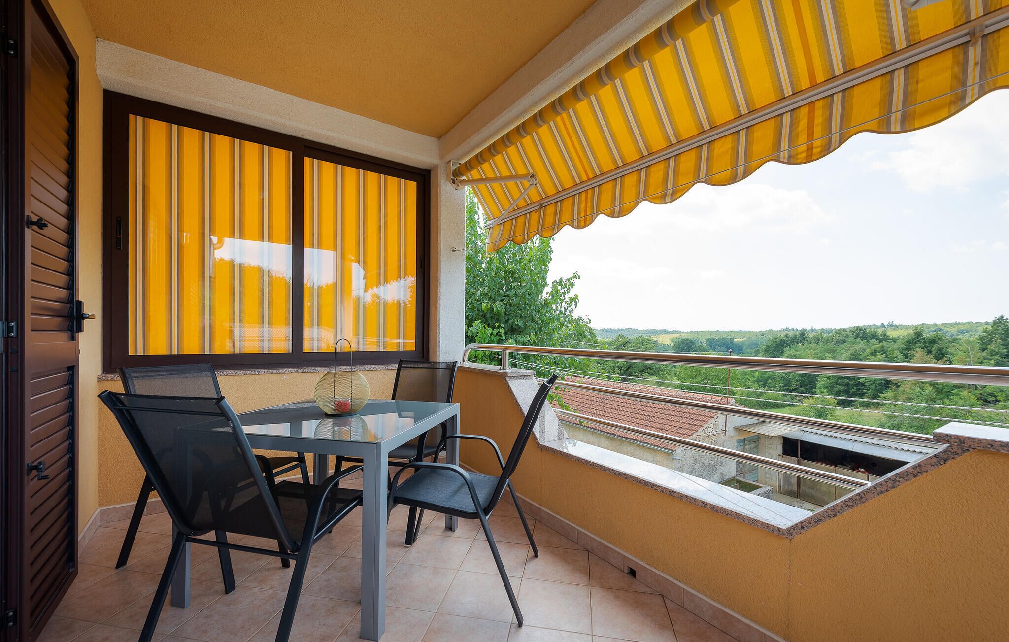 Een balkon met een tafel en stoelen, een geel zonnescherm en uitzicht op groen en gebouwen.