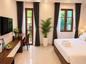 Habitación de hotel con cama, escritorio y silla, televisor, planta en maceta y ventana con cortinas.