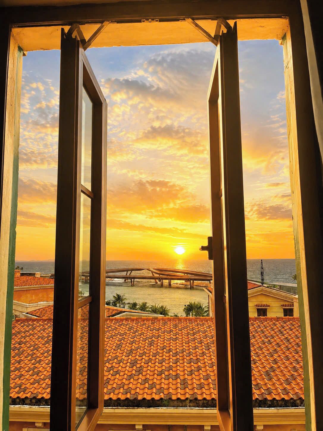 A view through an open window with a sunset over the ocean, a beach with a pier, and terracotta rooftops.