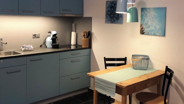A modern kitchen with blue cabinets, a coffee machine, a sink, and a dining table with chairs. - Mugliner 117A, (Ftan) by Interhome (Ftan)