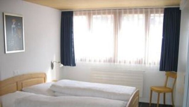Quarto com cama de casal, uma cadeira, radiador e janela com cortinas.