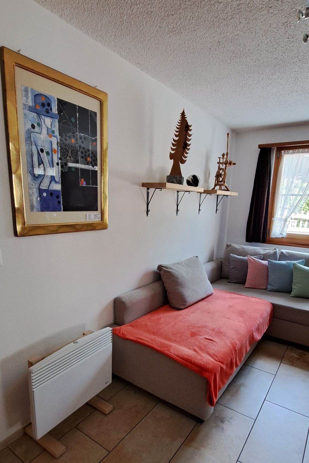Une pièce comprenant un canapé-lit, un radiateur, une fenêtre avec des rideaux et une étagère murale agrémentée d’objets décoratifs.