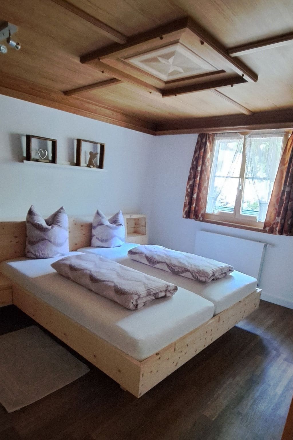 A bedroom with a wooden ceiling, two beds with pillows, a window with curtains, and a radiator.