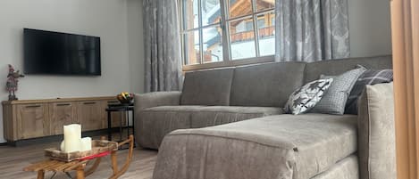 Uma sala de estar com um sofá cinzento, uma cadeira de balanço de madeira, uma televisão de tela plana e uma janela com cortinas.