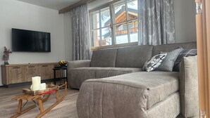 Un salón moderno con un sofá gris, una mecedora de madera, un televisor de pantalla plana y una ventana con cortinas.