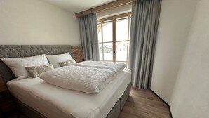 Una cama bien hecha con cabecera gris y ropa de cama blanca, una ventana con cortinas y piso de madera.