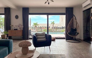 Ein modernes Wohnzimmer mit Blick auf ein Schwimmbad und Palmen, ein Hängesessel, eine Couch und ein Dekorationstisch.