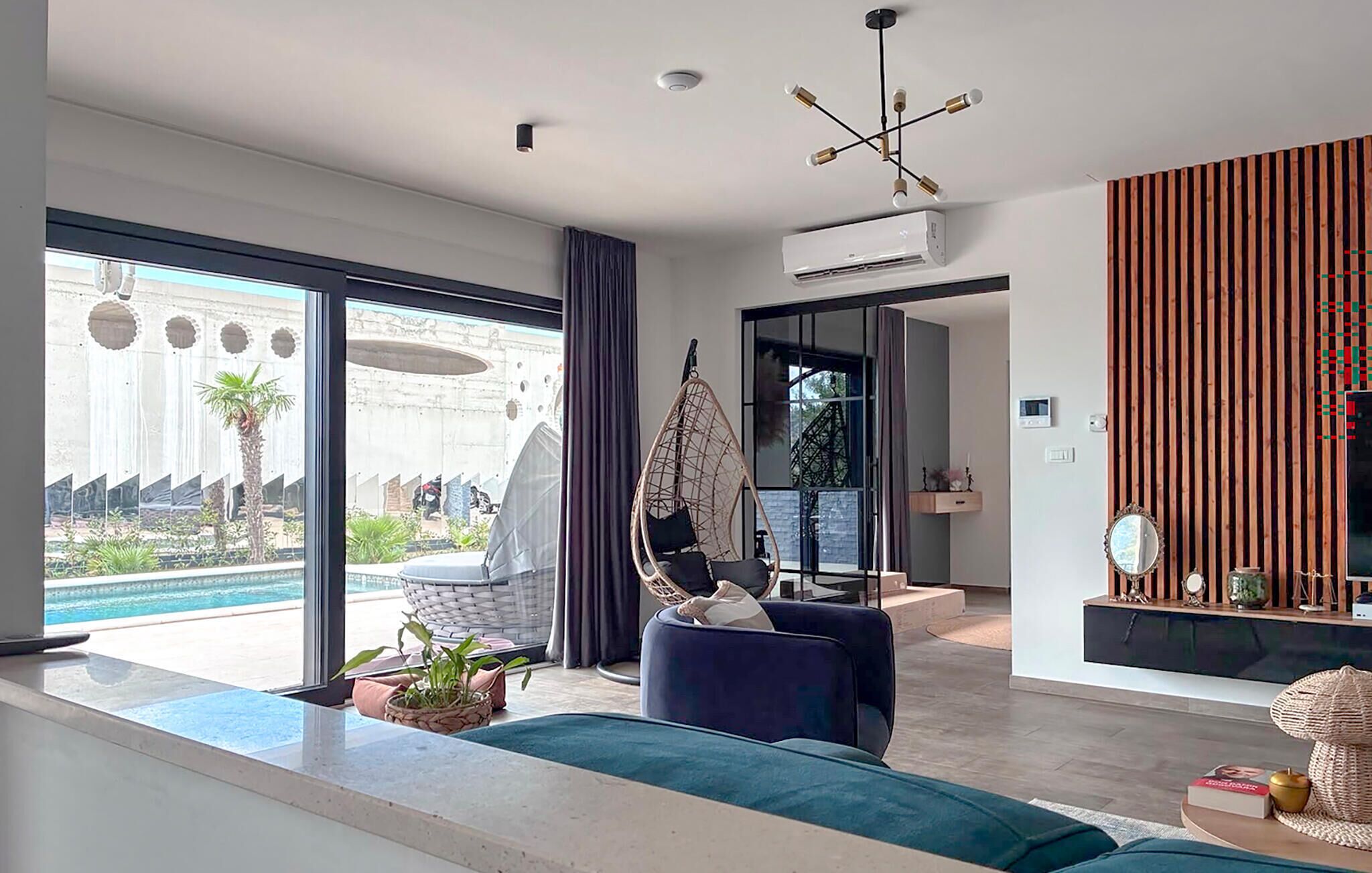 A modern living room with a large sliding glass door leading to a pool area, a hanging chair, and a wooden slatted wall.