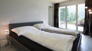Ein Doppelbett mit weißer Bettwäsche in einem modernen Hotelzimmer mit großem Fenster und Blick auf Grün.