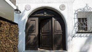 Una puerta de madera oscura con un arco blanco, una pared de piedra con leña apilada y una barandilla de metal para balcón.