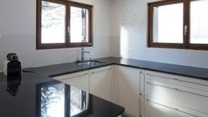 Une cuisine moderne avec des meubles blancs, un plan de travail noir et deux fenêtres à cadre en bois.