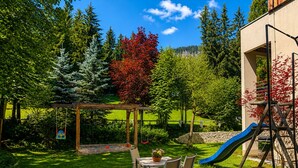 A backyard with a wooden swing set, a table with chairs, a slide, and a stone wall. - Mi Chalet (Badia)