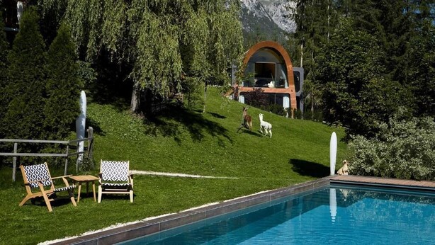 A poolside area with a view of a mountain, a wooden deck, and a building with a rounded archway. - Mi Chalet (Badia)
