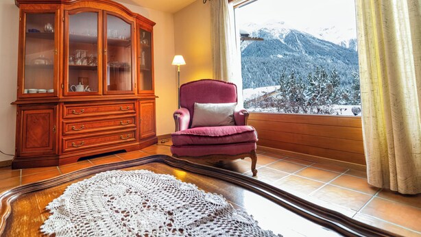 A room with a wooden cabinet, a red armchair, a view of snow-covered mountains, and a large window with curtains. - Ferienhaus Grundschera, (Brail) by Interhome (Brail)