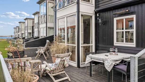 A wooden deck with outdoor seating, a table with a bottle and glasses, and a view of a building with large windows. - Nice home in Juelsminde with harbor view (Juelsminde)