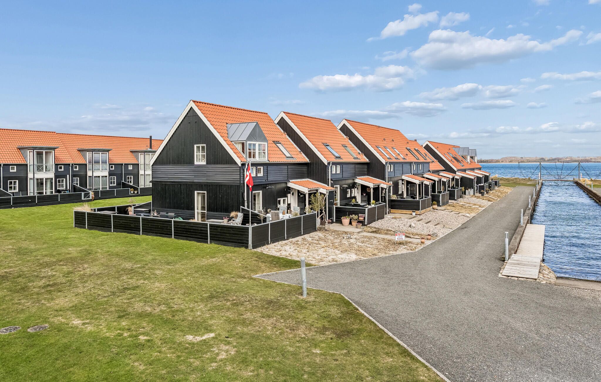 A row of modern, dark-colored houses with orange roofs, a grassy area, and a body of water in the background.