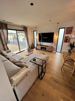 Ein modernes Wohnzimmer mit großem Flachbildfernseher, einer Couch, einem Couchtisch und einem Essbereich mit einem Stuhl.