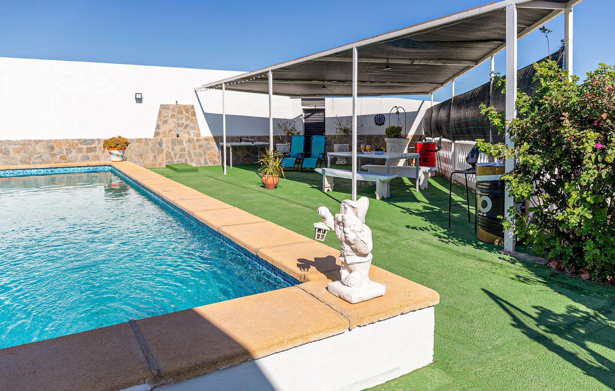 A swimming pool with a stone edge, a covered patio area with outdoor furniture, and a stone wall.