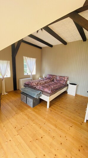 A bedroom with a wooden floor, a bed with patterned bedding, a bedside table, and a window with curtains. - Vacation at the Horse Farm—Pets Welcome! (Jade)
